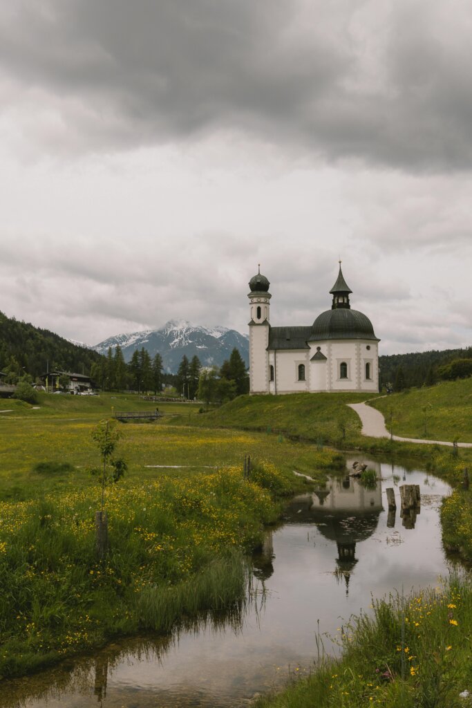 Picturesque church by a stream in a lush meadow with mountains behind.